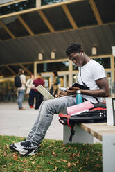 African male student reading book while sitting on bench in university campus - MASF22258