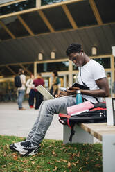 Afrikanischer männlicher Student liest ein Buch, während er auf einer Bank auf dem Universitätscampus sitzt - MASF22258