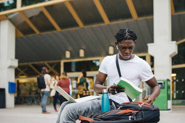 Männlicher Student, der ein Buch mit einem Laptop auf einem College-Campus hält - MASF22260