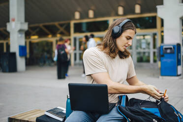 Männlicher Student mit Laptop auf einer Bank auf dem Campus sitzend - MASF22266