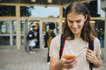 Lächelnder männlicher Student, der ein Mobiltelefon in der Universität benutzt - MASF22270