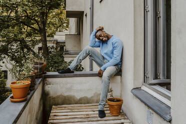 Lächelnder Mann sitzt mit dem Kopf in der Hand auf einer Stützmauer am Balkon - MASF22279