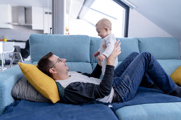 Mid adult man wearing eyeglasses lying on sofa while playing with son at home - AMPF00099