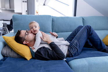 Affectionate father kissing baby son while lying on sofa in living room - AMPF00100