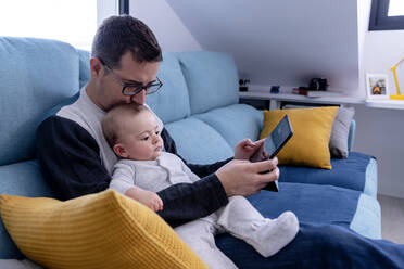 Father kissing son's head while sitting on sofa with tablet at home - AMPF00102
