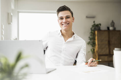 Smiling young man with laptop sitting at desk - UUF22821