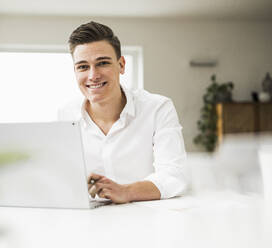 Lächelnde männliche Fachkraft, die einen Laptop benutzt, während sie am Tisch im Heimbüro sitzt - UUF22824