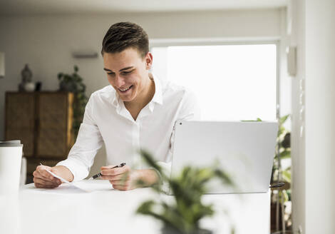Happy young businessman reading document while sitting in home office - UUF22825