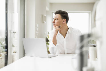 Junger männlicher Berufstätiger, der durch das Fenster schaut, während er in seinem Büro zu Hause sitzt - UUF22827