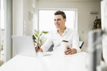 Lächelnder Geschäftsmann mit Einweg-Kaffeebecher und Laptop am Schreibtisch sitzend - UUF22828