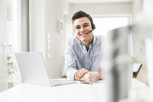 Young male entrepreneur wearing headset while sitting at home office - UUF22832