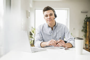 Male professional wearing headset sitting with laptop at desk - UUF22833