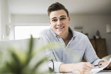 Lächelnder junger Geschäftsmann mit Dokument am Tisch sitzend im Heimbüro - UUF22840