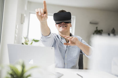 Lächelnder männlicher Berufstätiger, der einen Virtual-Reality-Simulator trägt und gestikuliert, während er im Büro zu Hause sitzt - UUF22842