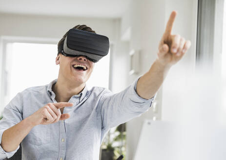 Cheerful male entrepreneur gesturing while wearing virtual reality simulator at home office - UUF22844