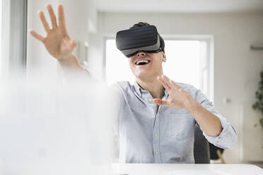 Cheerful male professional wearing wearing virtual reality simulator while sitting at desk in home office - UUF22845