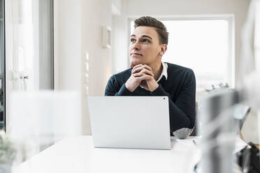 Thoughtful businessman with laptop looking away while sitting at desk - UUF22846