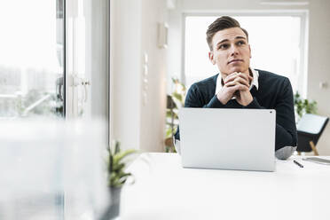 Nachdenklicher männlicher Unternehmer mit Laptop, der am Schreibtisch sitzt und wegschaut - UUF22847