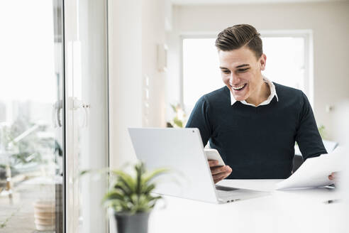 Fröhlicher Geschäftsmann, der eine SMS auf seinem Smartphone schreibt, während er im Home Office sitzt - UUF22852