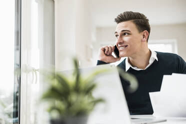 Lächelnder männlicher Unternehmer, der am Telefon spricht und durch ein Fenster im Büro zu Hause schaut - UUF22856