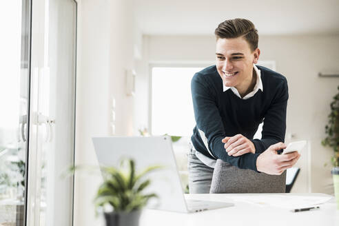 Happy young businessman leaning on chair while looking at laptop in home office - UUF22858