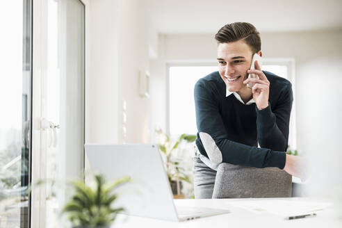 Männlicher Unternehmer, der am Telefon spricht, während er sich im Büro zu Hause an einen Stuhl lehnt - UUF22859