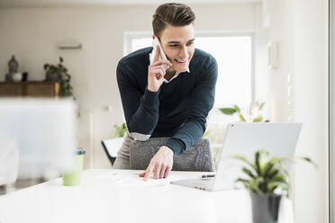 Lächelnder männlicher Unternehmer, der mit seinem Handy telefoniert, während er sich im Büro zu Hause an einen Stuhl lehnt - UUF22861