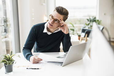 Thoughtful male entrepreneur with document sitting at desk looking away - UUF22867