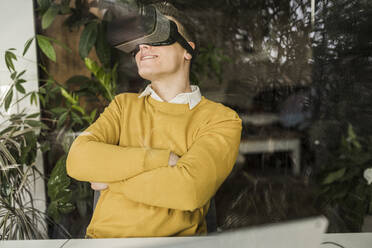 Lächelnder männlicher Unternehmer, der einen Virtual-Reality-Simulator trägt, gesehen durch ein Fenster im Heimbüro - UUF22891
