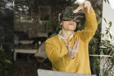 Young businessman gesturing while wearing virtual reality simulator seen through window in home office - UUF22893