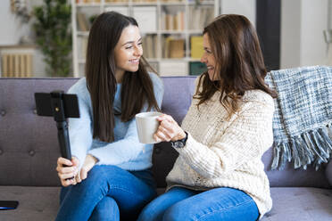 Smiling daughter holding mobile phone through monopod while talking with mother on sofa at home - GIOF11422