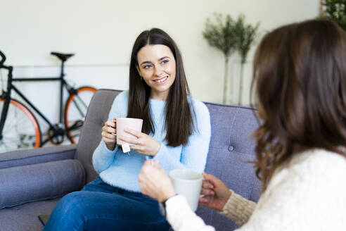 Lächelnde Tochter mit Kaffeetasse im Gespräch mit der Mutter auf dem Sofa zu Hause - GIOF11424