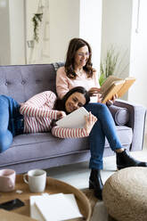Lächelnde Tochter, die ein digitales Tablet benutzt, während sie auf dem Schoß der Mutter liegt und ein Buch auf dem Sofa zu Hause liest - GIOF11428