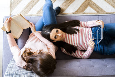 Daughter with digital tablet lying on lap of mother holding book on sofa - GIOF11431