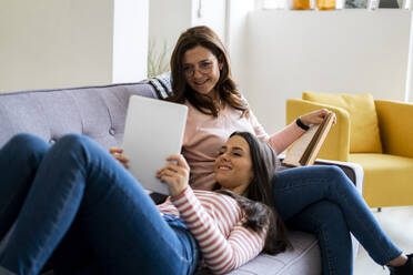 Smiling daughter showing digital tablet while lying on lap of mother with book sitting on sofa at home - GIOF11432