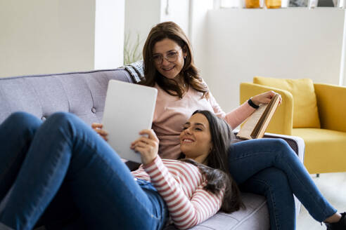 Lächelnde Tochter zeigt digitales Tablet, während sie auf dem Schoß der Mutter mit einem Buch auf dem Sofa zu Hause sitzt - GIOF11432