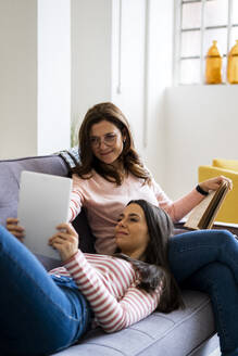 Lächelnde Mutter und Tochter schauen gemeinsam auf dem Sofa zu Hause auf ein digitales Tablet - GIOF11433