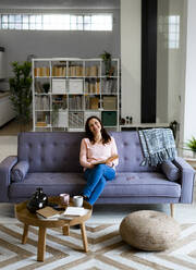 Lächelnde reife Frau mit Buch sitzt auf dem Sofa zu Hause - GIOF11434