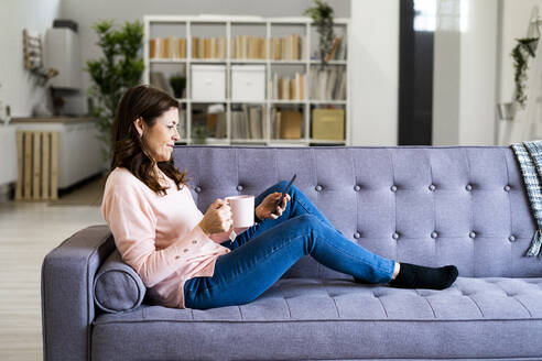 Frau mit Kaffeebecher und Mobiltelefon, während sie zu Hause auf dem Sofa sitzt - GIOF11438