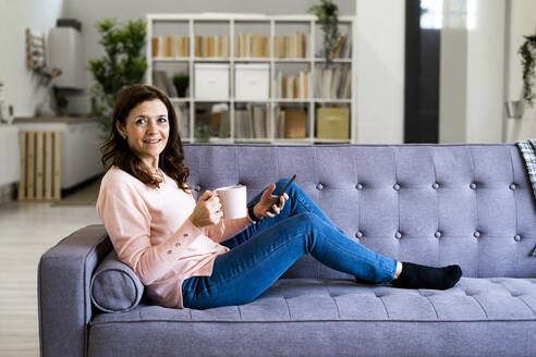 Lächelnde Frau mit Kaffeebecher in der Hand, während sie zu Hause auf dem Sofa sitzt - GIOF11439