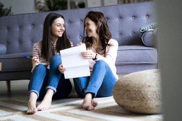 Mother with document smiling at daughter while sitting on floor in living room - GIOF11443