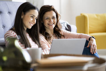 Smiling mother and daughter looking at laptop together while sitting in living room at home - GIOF11448