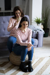 Smiling mother combing daughter's hair at sofa in living room - GIOF11455