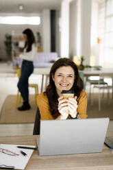 Smiling businesswoman holding coffee cup while working at home - GIOF11463