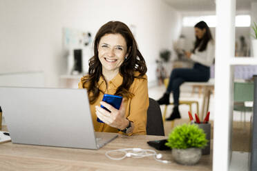 Lächelnde Geschäftsfrau mit Laptop, die ihr Smartphone hält, während sie an einem Schreibtisch im Heimbüro sitzt - GIOF11464