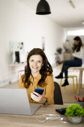 Lächelnde Geschäftsfrau mit Laptop, die ihr Smartphone hält, während sie an einem Schreibtisch im Heimbüro sitzt - GIOF11465