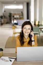 Happy businesswoman with laptop looking away while talking on mobile phone at home office - GIOF11467