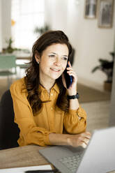 Lächelnde Geschäftsfrau mit Laptop, die auf einem Smartphone spricht, während sie am Schreibtisch im Heimbüro sitzt - GIOF11468