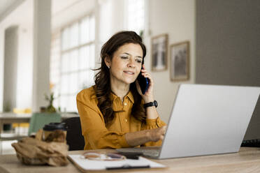 Lächelnde Geschäftsfrau, die einen Laptop benutzt, während sie am Schreibtisch im Heimbüro mit dem Handy telefoniert - GIOF11469