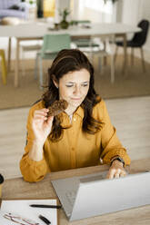Geschäftsfrau, die einen Keks isst, während sie einen Laptop am Schreibtisch im Heimbüro benutzt - GIOF11470